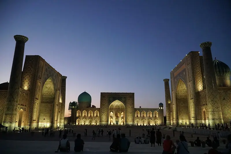 Registan Square in Samarkand at dusk, its three madrasas lit gold against a deep blue sky.