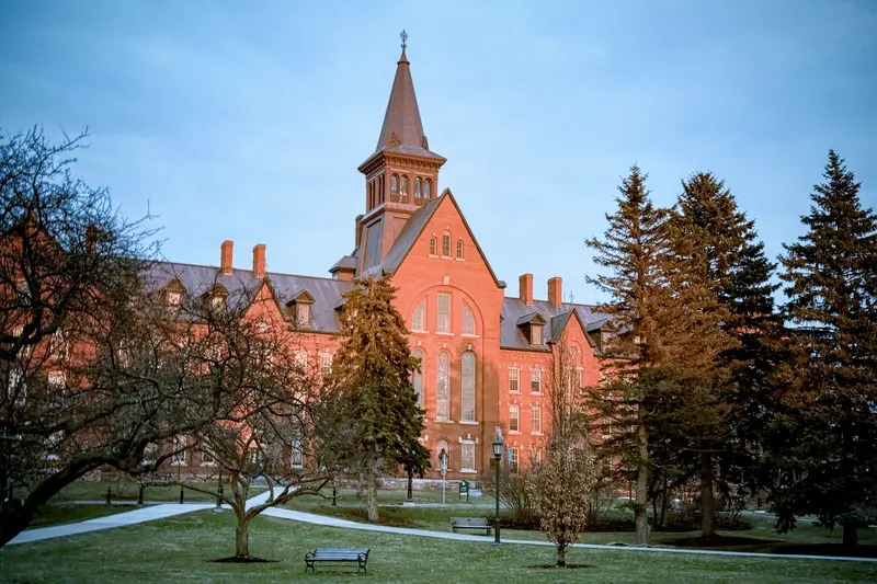 The Old Mill at the University of Vermont in Burlington, a long red-brick building with a tall spire, among evergreens.